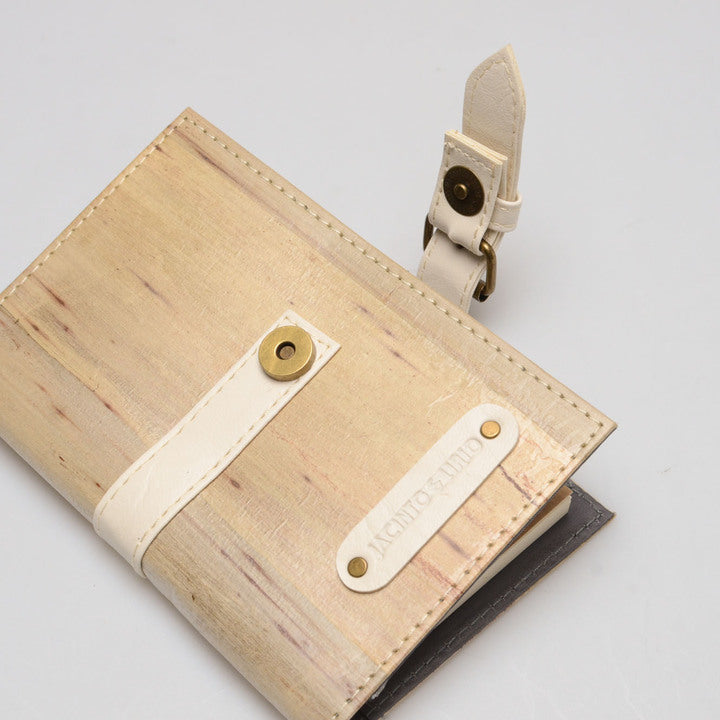 A pale beige vegan leather journal lying on a surface, partially opened with its white belt buckle closure unclasped.