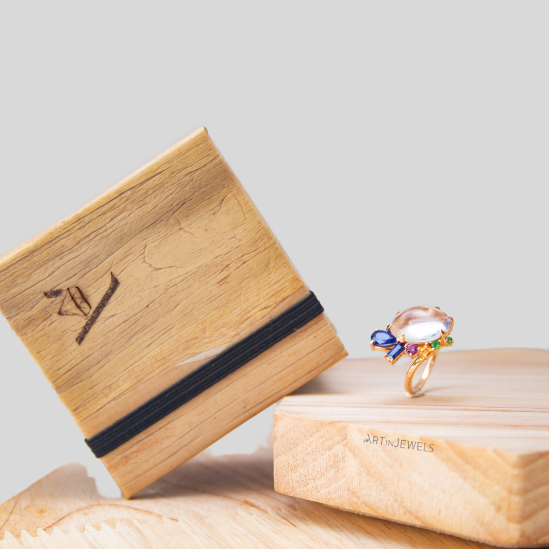 A colorful multi-gemstone ring sitting on top of a pale wooden platform, with a simplistic jewelry box with a wooden aesthetic and a black garter leaning on the wooden platform diagonally.