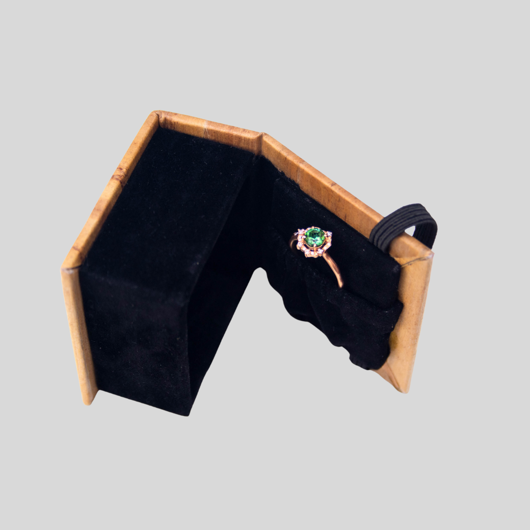 A black ring box with a vegan leather exterior and black velvet fabric interior laying on its side, with an emerald ring resting in a garterized compartment on the inner side of the lid of the box.