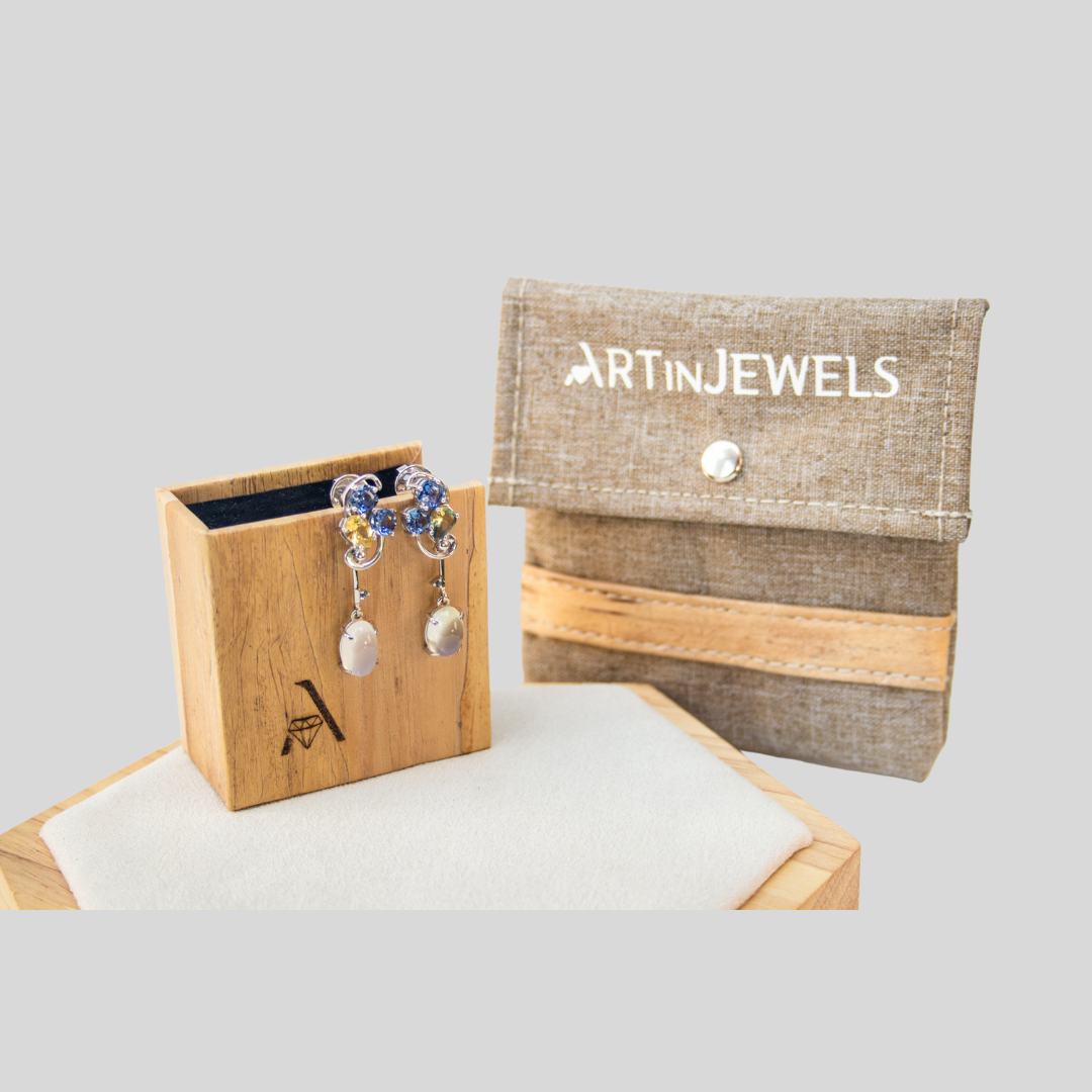 A pair of dangling earrings hanging from the side of a vegan leather jewelry box, next to a fabric bag with a horizontal vegan leather strip for accent.