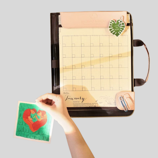 A hand holding up an abstract illustration depicting love, next to a vegan leather dated planner with a handle on its side.