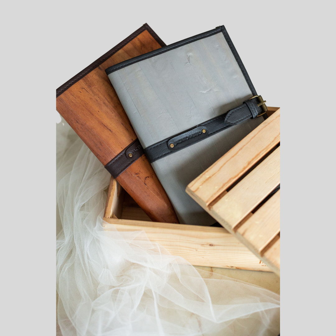 Two vegan leather notebooks peeking out of a pale wooden crate, one with a dark brown cover resembling wood and the other pale blue with a thin belt buckle closure.