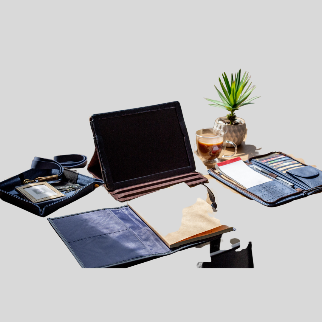 A desktop of stylish work items all made of vegan leather, such as a catch all tray, a blue refillable notebook, a vegan leather ipad case, a wallet containing business cards and a checkbook, and some coffee.