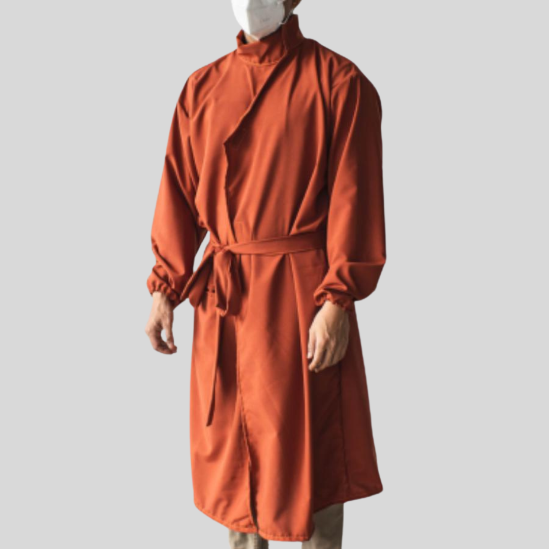 A masked individual wearing an orange PPE suit that has a stylish v-neck collar and built-in belt that can be tied for closure, giving it the feel of a coat.