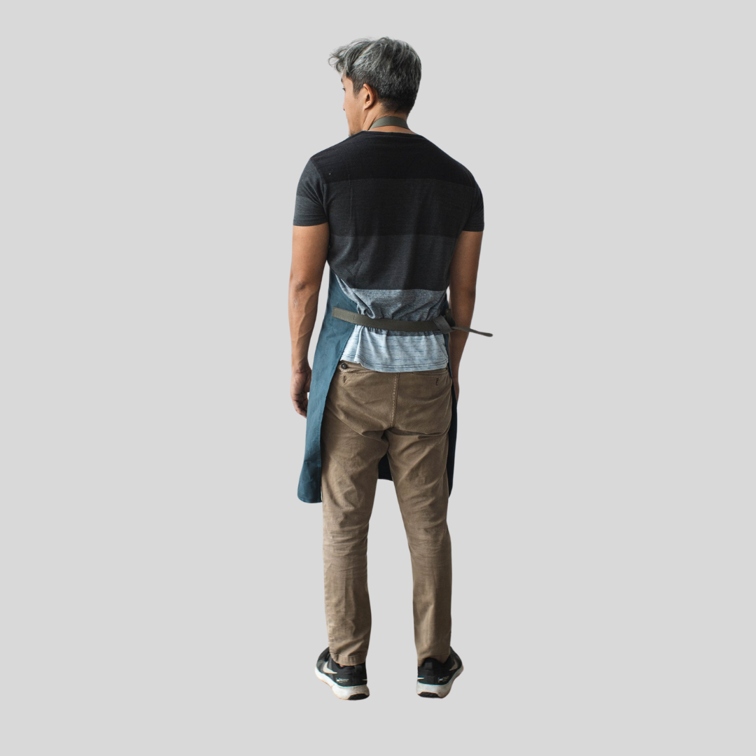 A man standing upright and showcasing the back of an apron that has one strap that wraps around his waist and another that goes around his neck.