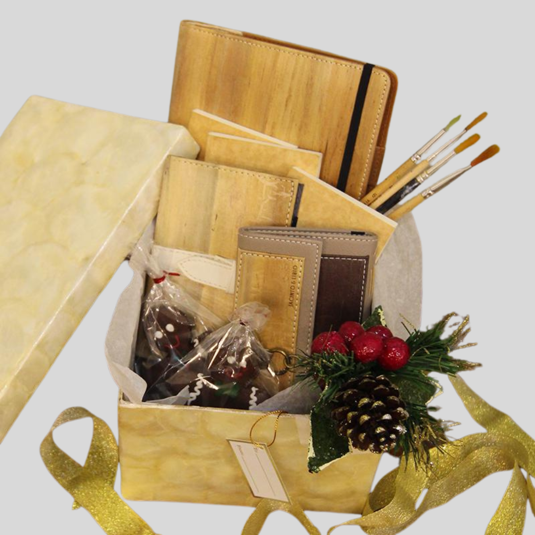 Beige box containing products made of vegan leather such as notebooks, wallets, an ipad case, and some paintbrushes. A gold ribbon lays around it while a pine cone is pinned on the box.