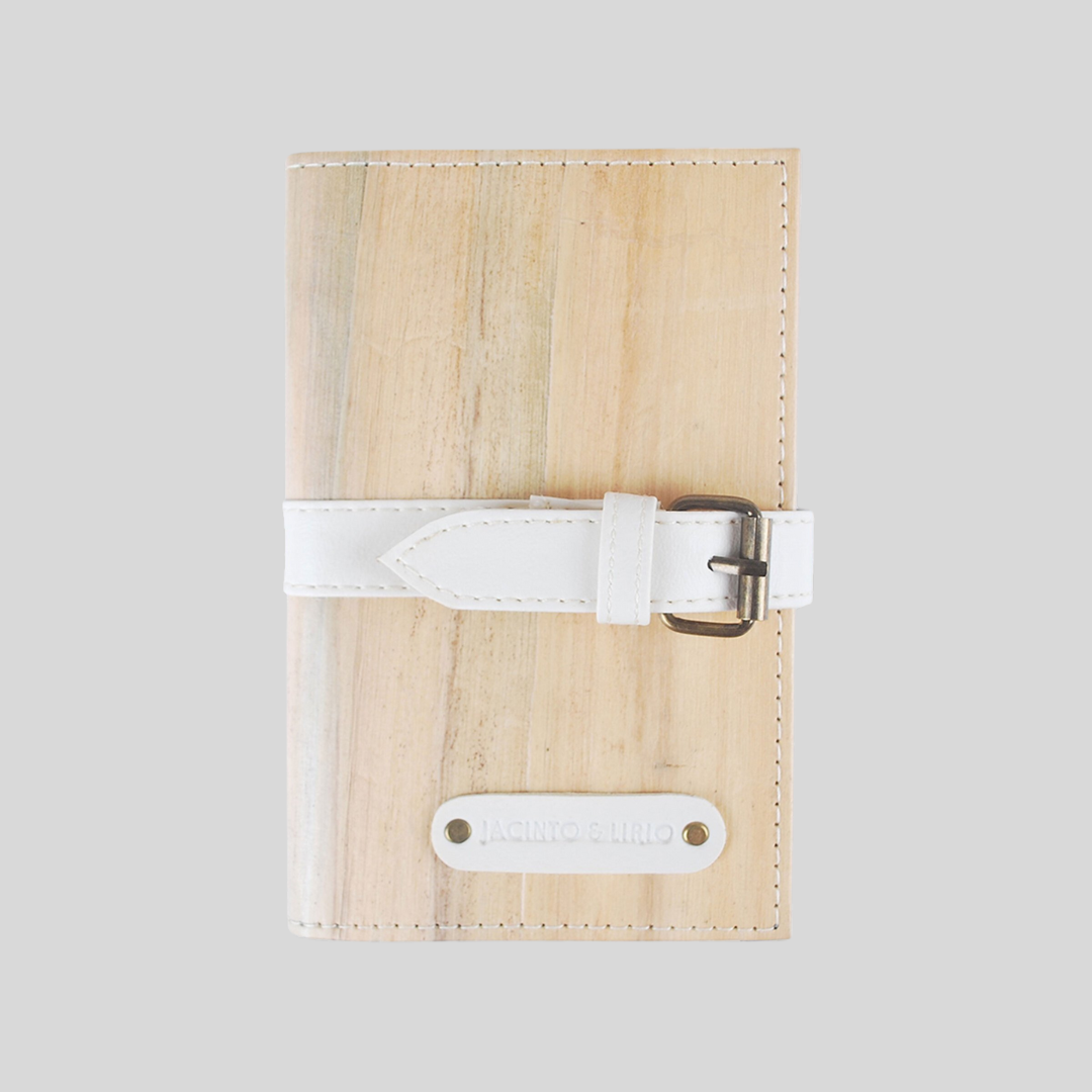 A notebook made of pale vegan leather from water hyacinth stalks, closed shut by a white leather belt buckle going around its body.