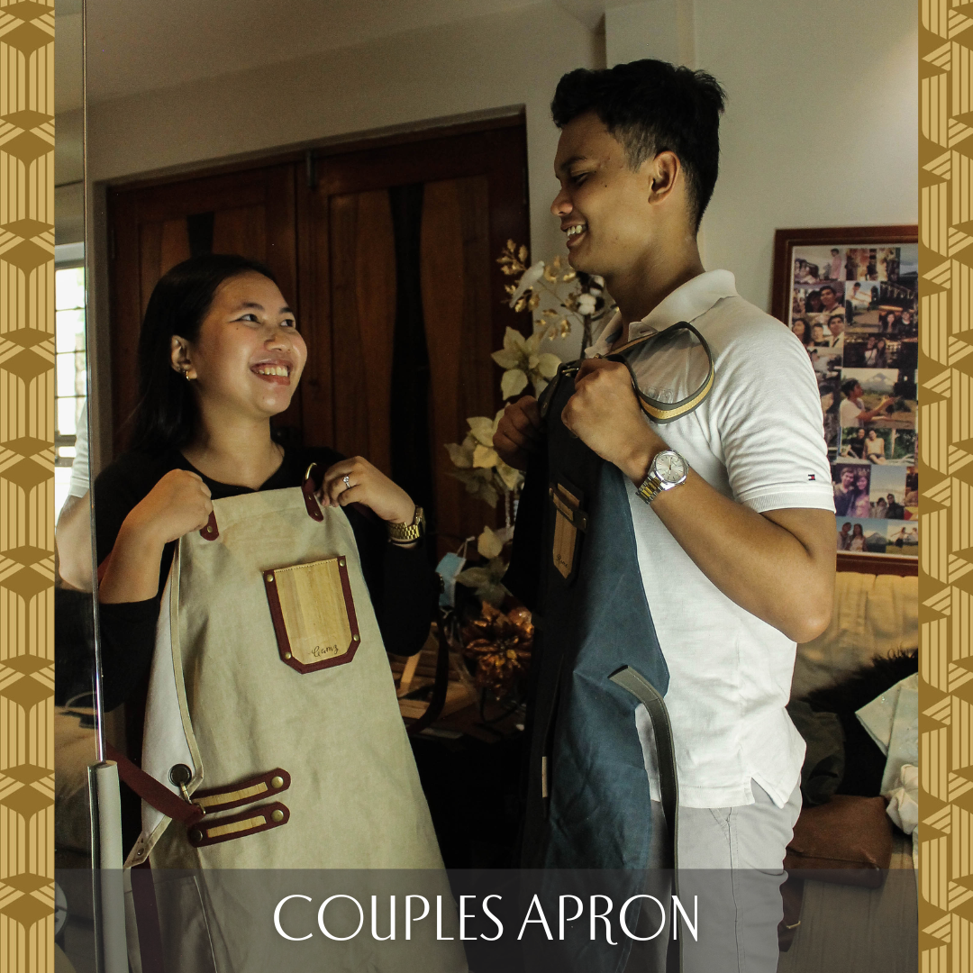 A couple looking at each other and holding up matching aprons, the woman holding a biege apron and the man holding a dark blue one.