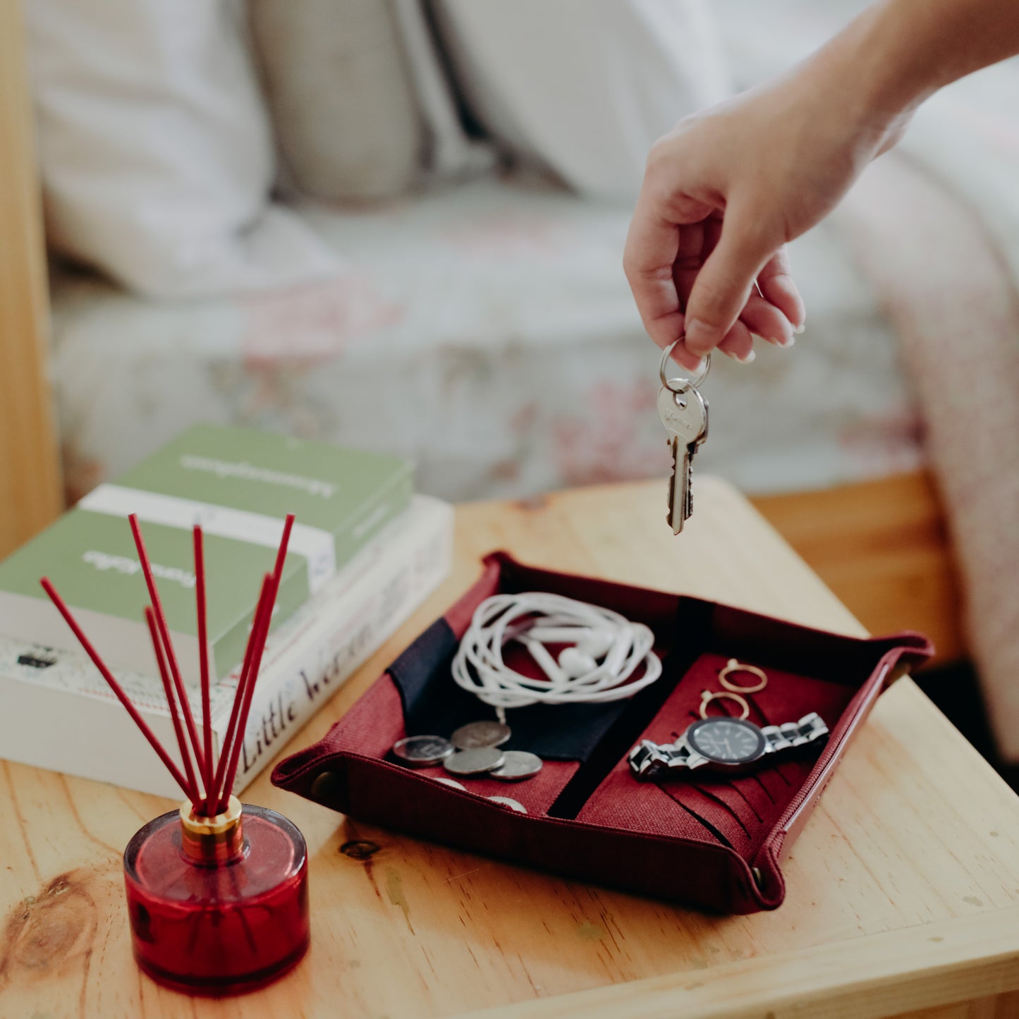 A key about to be dropped onto a dark red vegan leather tray that contains bedside items such as earphones, jewelry, and coins.