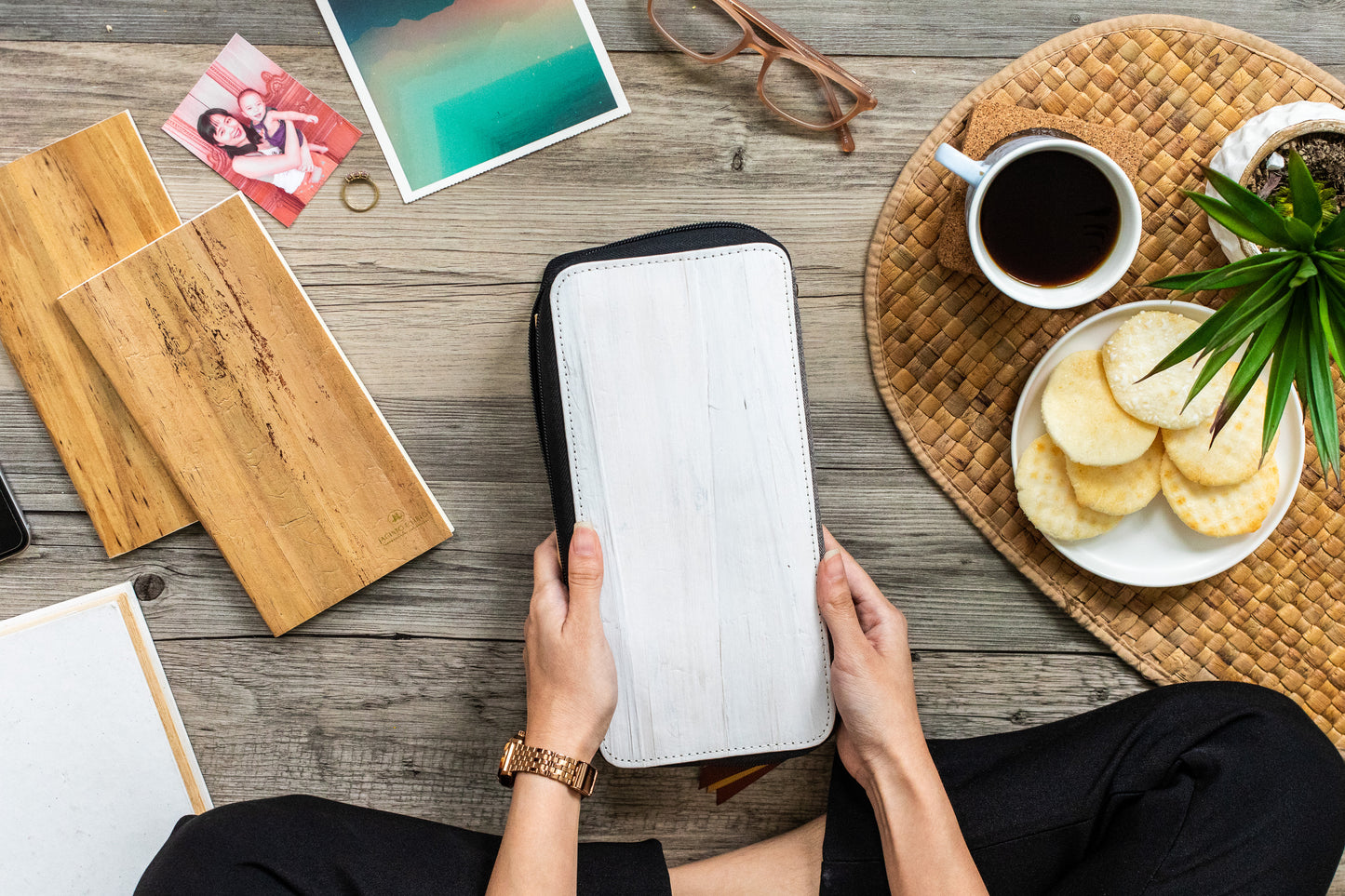 A person sitting cross legged on the ground beside some notebooks, a tray of biscuits and coffee, and holding in their hand a white multifunction rectangular notebook and wallet.