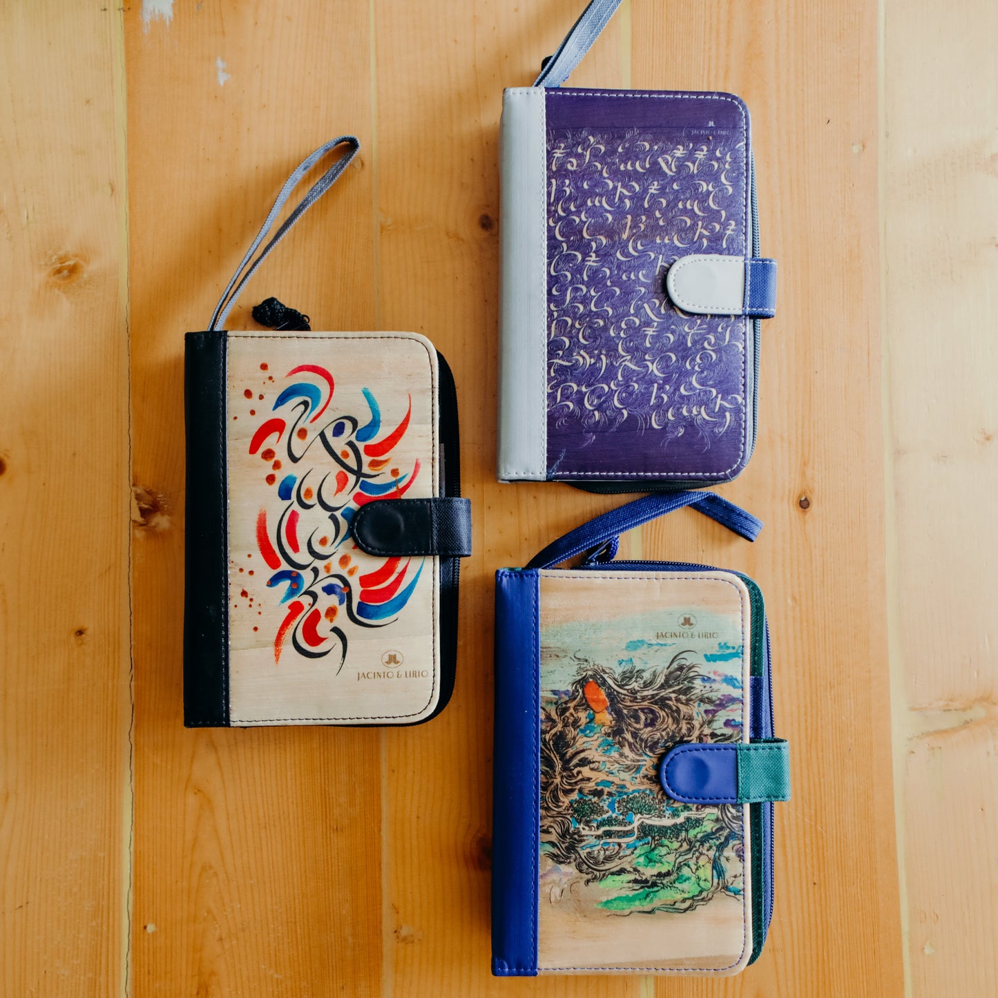 3 differently designed and colored vegan leather planners laying on a wooden surface, one violet and gray, the other beige and black, and the other blue and beige. All with hand-painted cover arts.