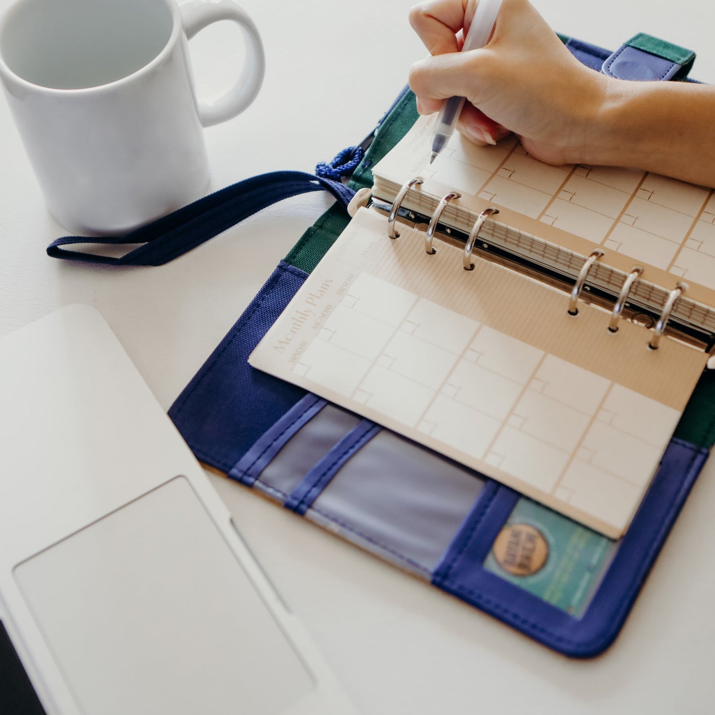 A person writing on a blue vegan leather dated planner with a cup of coffee and a macbook to the side.