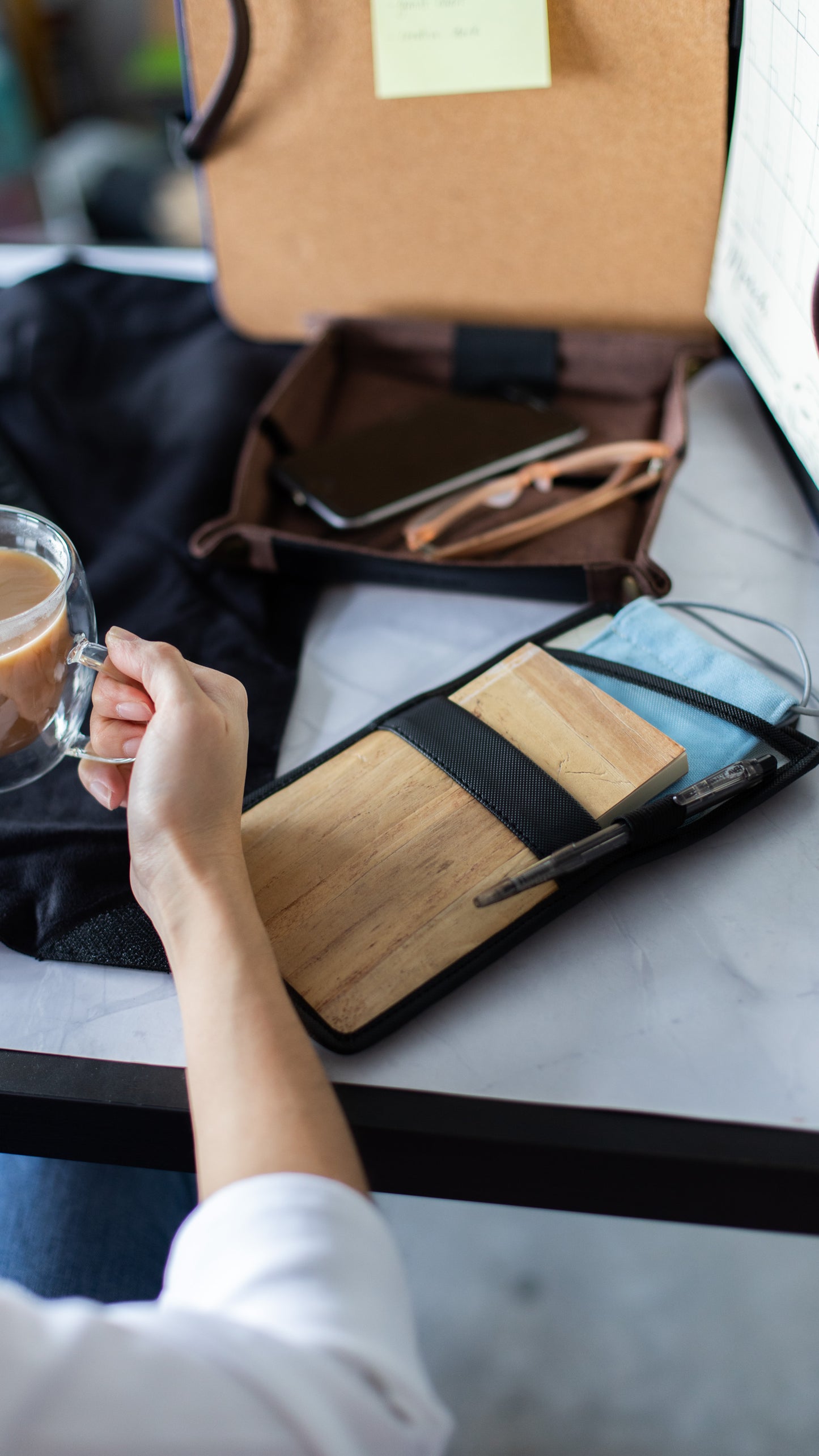 A person sipping coffee at their desk while a vegan leather notebook holder holds their notepad and pen.