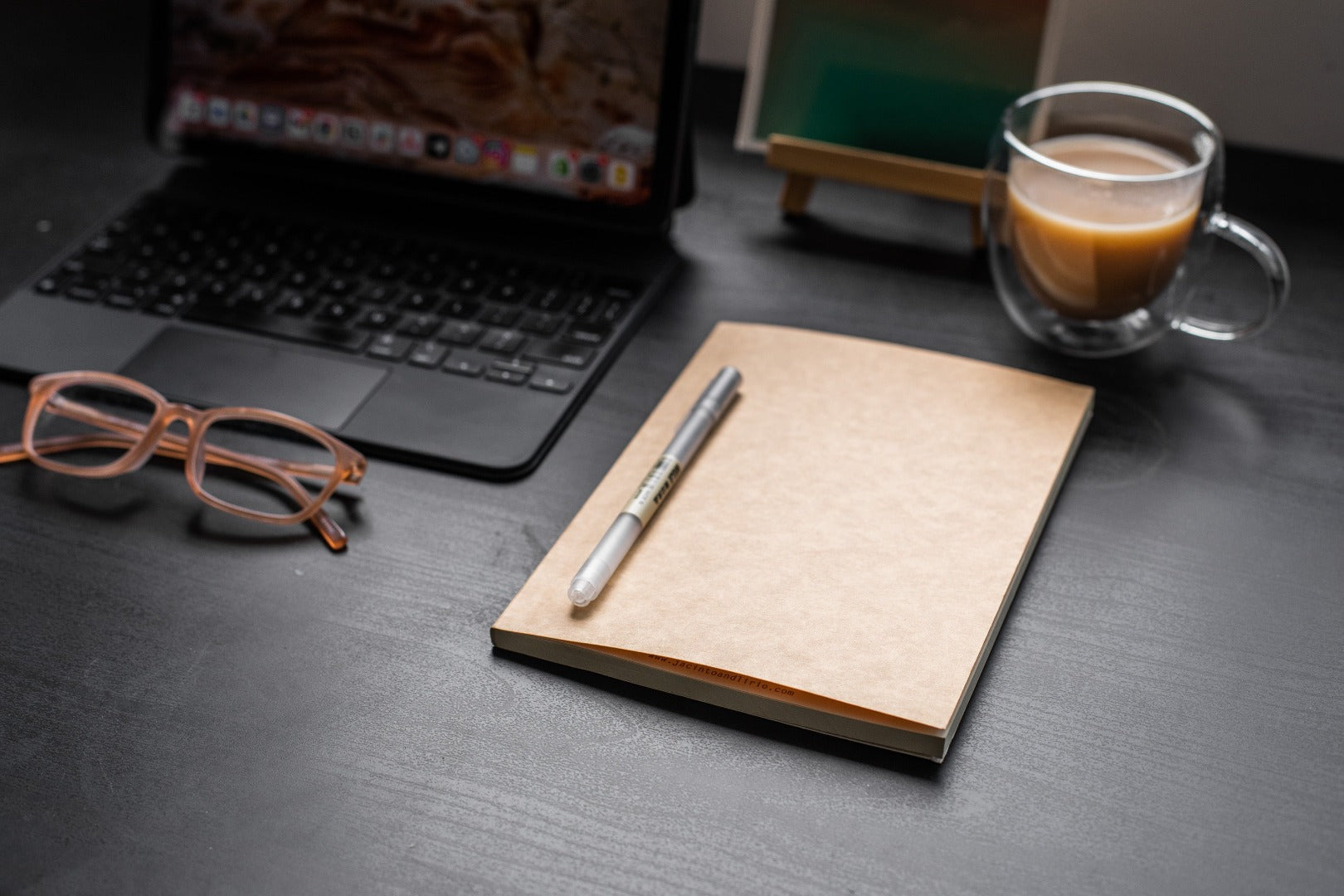 A black desktop with glasses, an ipad with a keyboard, a mug filled with coffee,  a closed medium sized notebook refill, and a pen, atop it.