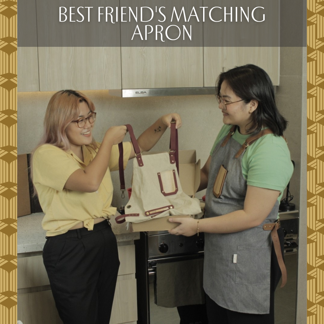One woman wearing a green shirt and gray apron joyfully handing another woman wearing a yellow shirt a matching cream-colored apron.