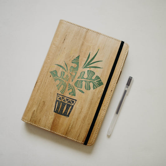 A wood-textured notebook that has a minimalist stamped design of a plant on its cover, the notebook has a black garter closure and beside it lays a Muji pen.