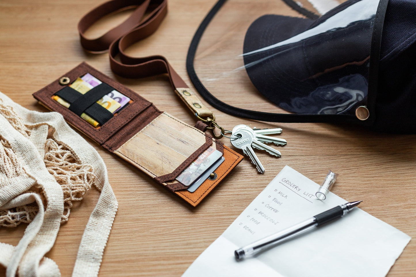 A brown vegan leather ID lanyard with cards, bills, and keys attached sprawled on a wooden surface next to a bag, face mask, piece of paper, and pen.