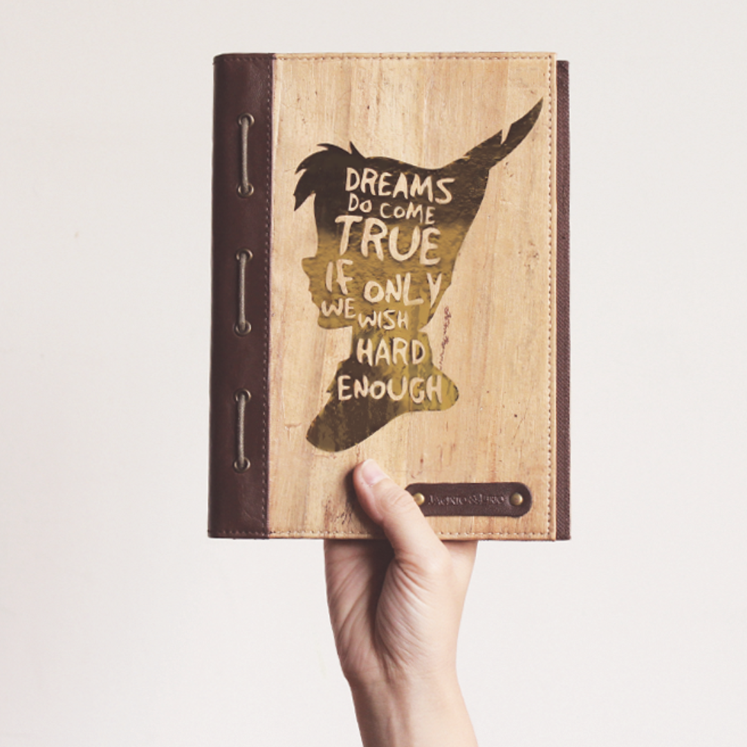 A hand holding up a vegan leather journal with an illustration of peter pan and a quote on it
