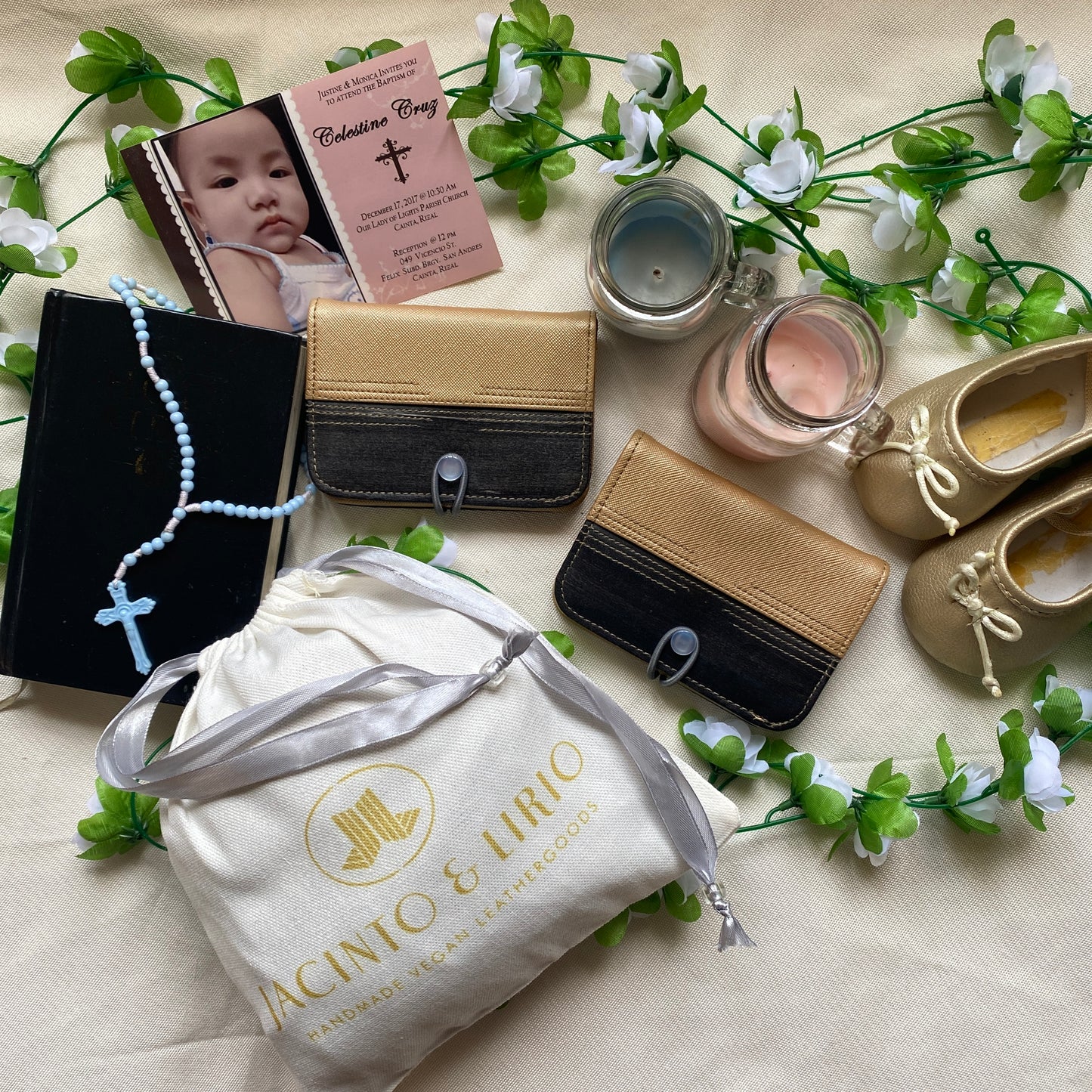 Baby shower goods organized aesthetically on a white tablecloth, such as sparkly baby shoes, some candles, two tiny vegan leather wallets, a cloth drawstring bag, a Bible and rosary, and a picture of the baby.