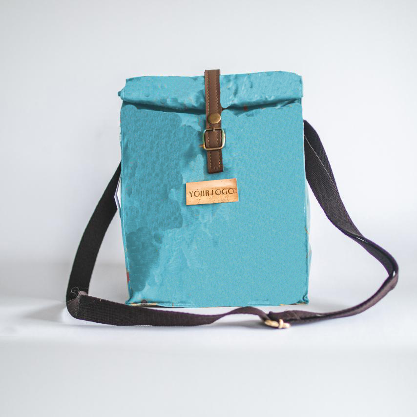 A light, sky blue lunch bag with a water-repellant waxed canvas exterior, vegan leather name patch, and a black strap.