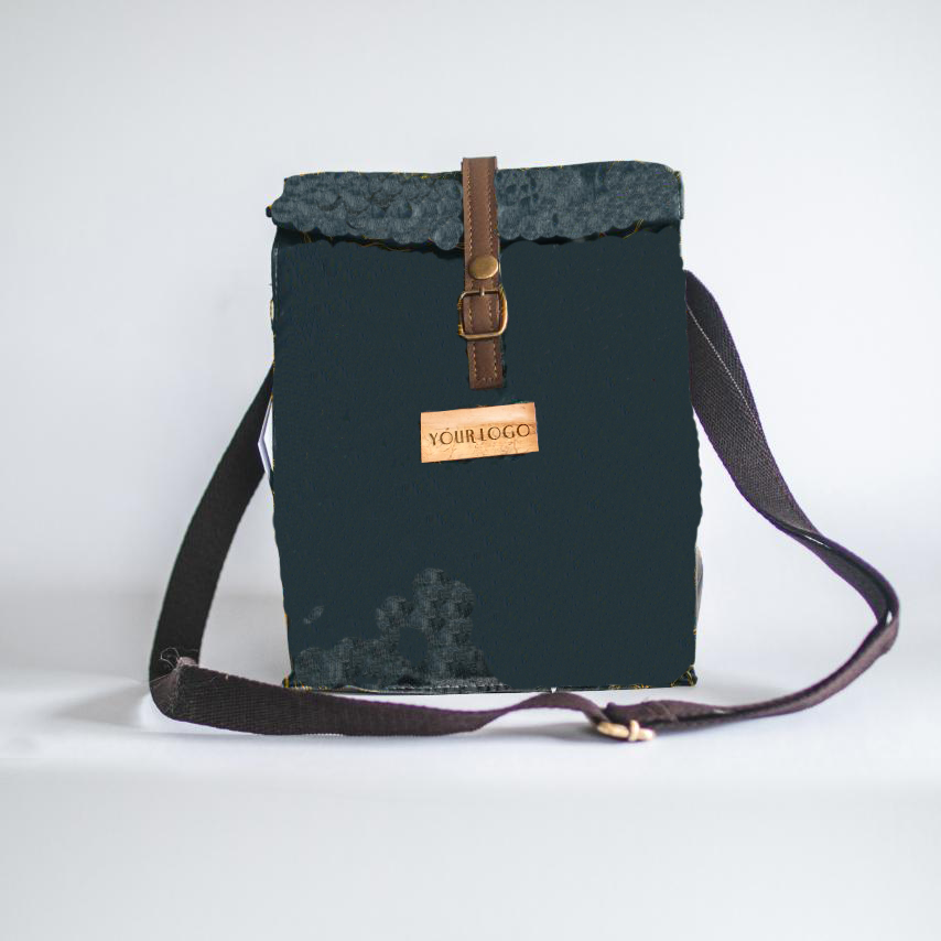 A black water-repellant lunch bag with a brown closing strap and a vegan leather logo patch.
