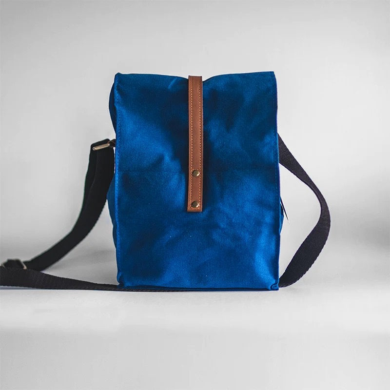 The back of a royal blue waxed canvas lunch bag, showing a brown vegan leather closing strap and a black cross-body strap.