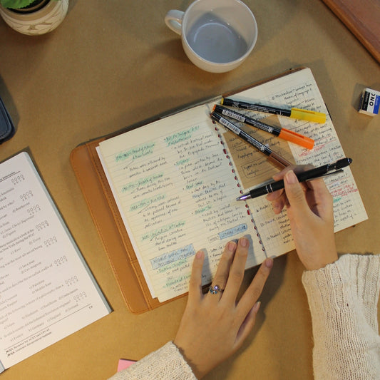 Person writing in a notebook in a Makata II Medium sleeve with markers on top, next to a mug and another open book.