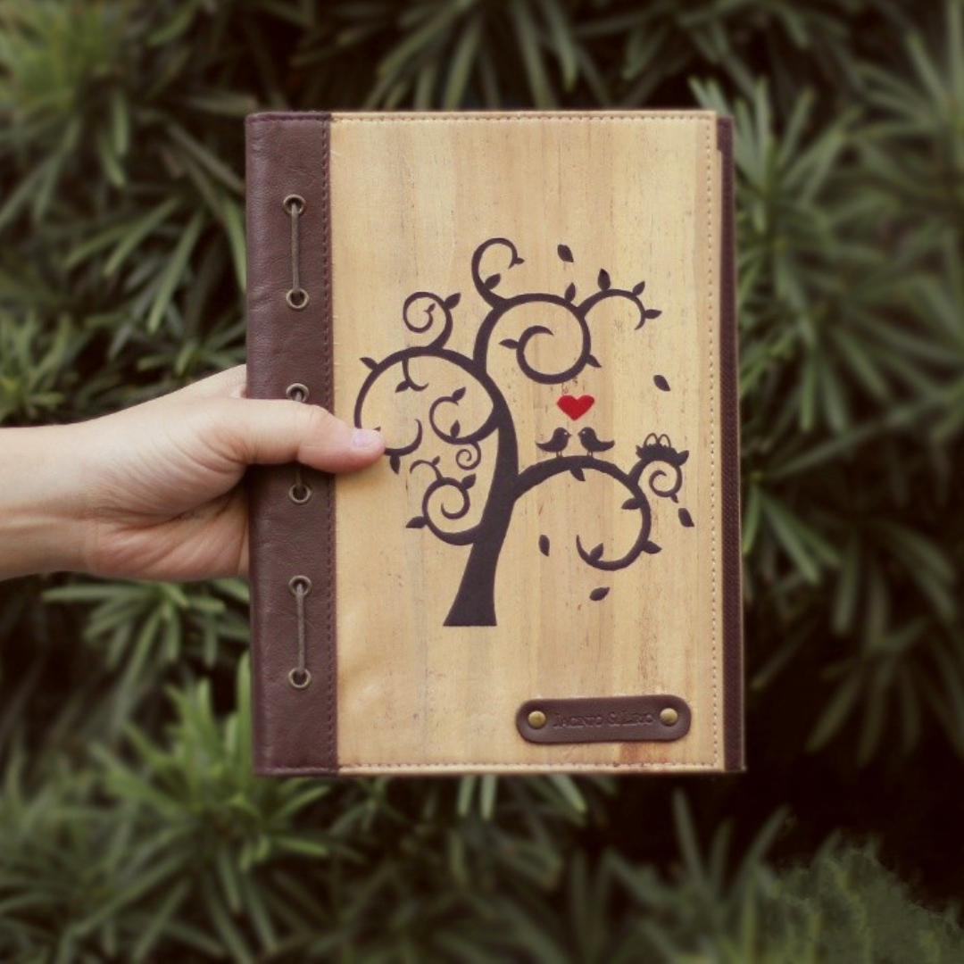A hand holding up a leather journal that has a drawing of a tree with two lovebirds on it.