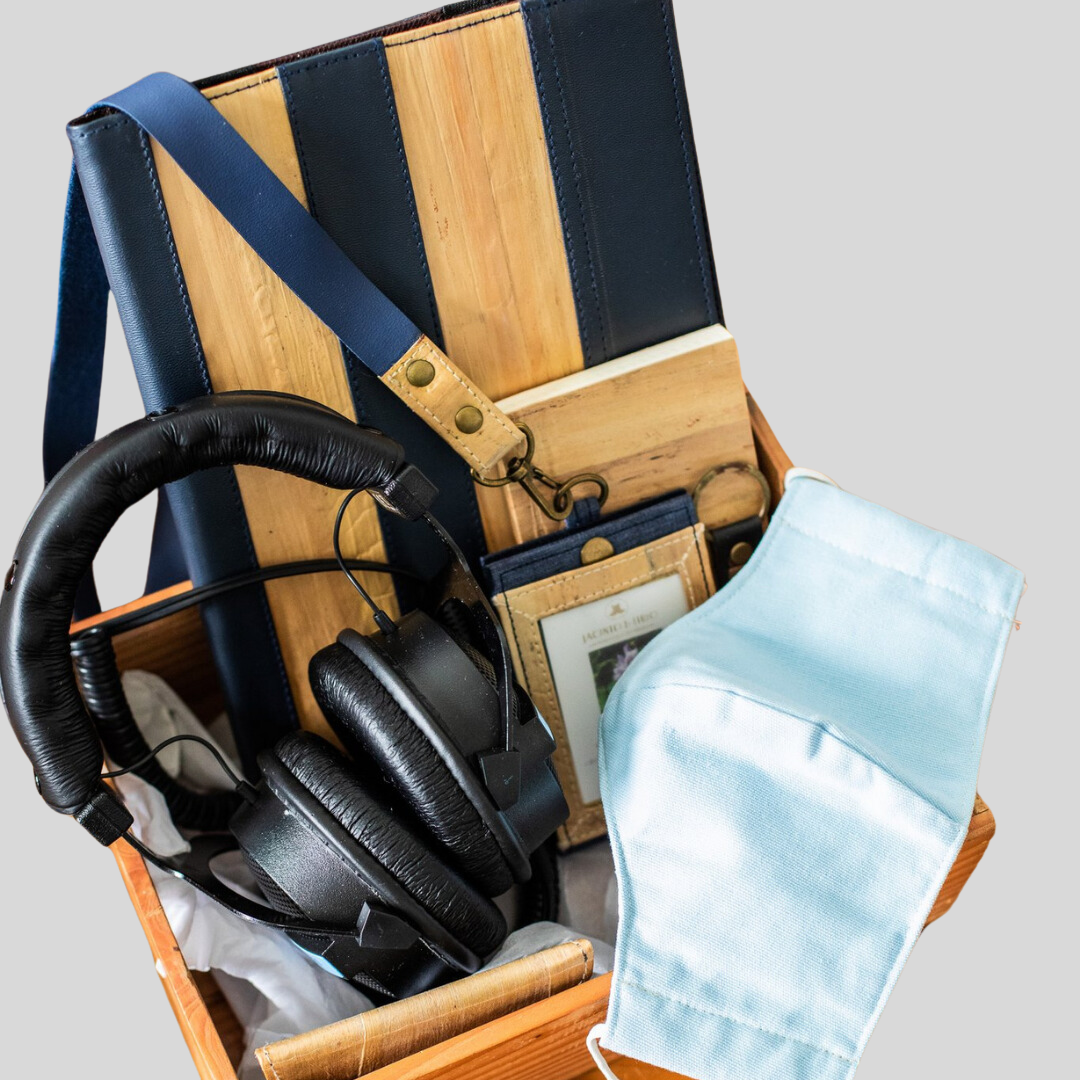 A wooden crate containing black headphones, a fabric surgical mask, a blue vegan leather ID lanyard, a small notebook, and a vegan leather iPad case.