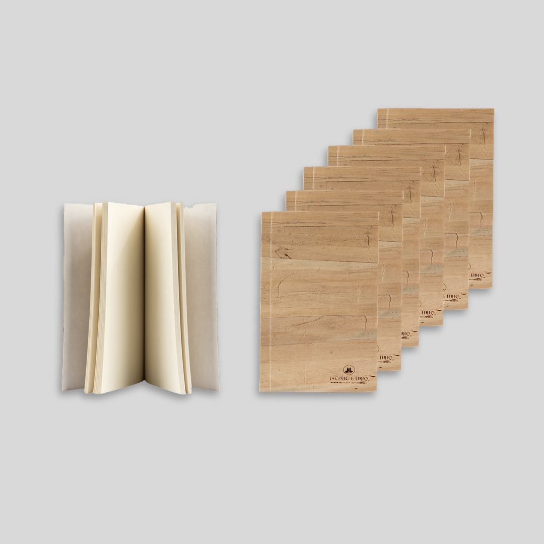 A notebook standing upright, partially opened showing blank pages while 6 closed notebooks with water hyacinth leather exteriors are stacked beside it.