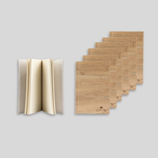 A notebook standing upright, partially opened showing blank pages while 6 closed notebooks with water hyacinth leather exteriors are stacked beside it.