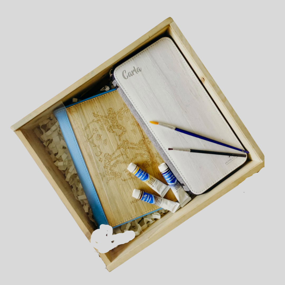 A wooden box with two vegan leather journals lying inside as well as some paintbrushes and tubes of paint.