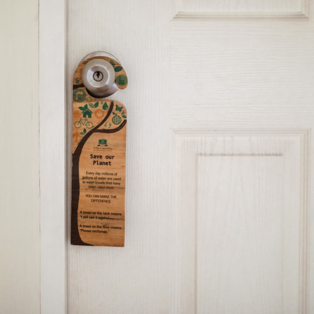 Eco-friendly hotel door hanger crafted from sustainable water hyacinth vegan leather, displaying a "Save our Planet" message with green environmental icons. The signage, promoting towel reuse and water conservation, hangs on a white hotel room door handle, ideal for green tourism initiatives.