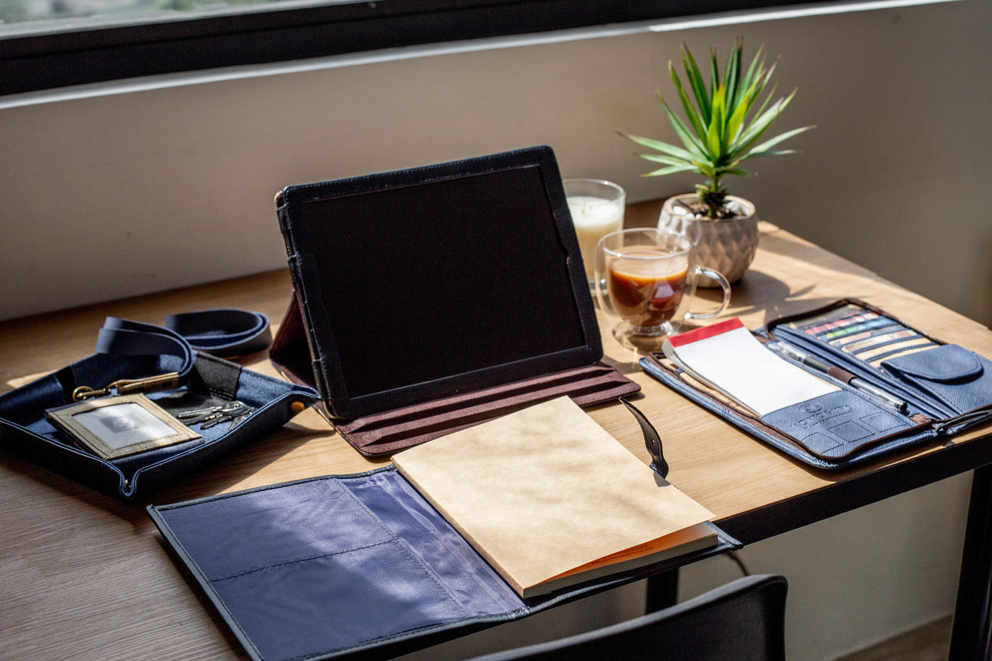 A sunny workspace with matching vegan leather items in navy blue, such as a valet tray containing an ID lanyard, a refillable notebook, a notepad holder with business cards, and an IPad case.