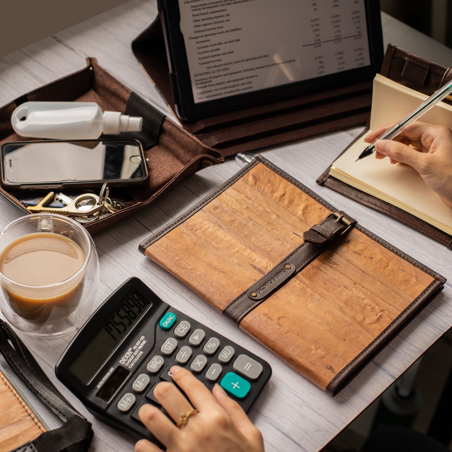 A person doing calculations on an aesthetic workplace featuring brown, earth-toned vegan leather goods such as a brown vegan leather ipad case, valet tray, and refillable cream-paged notebook.