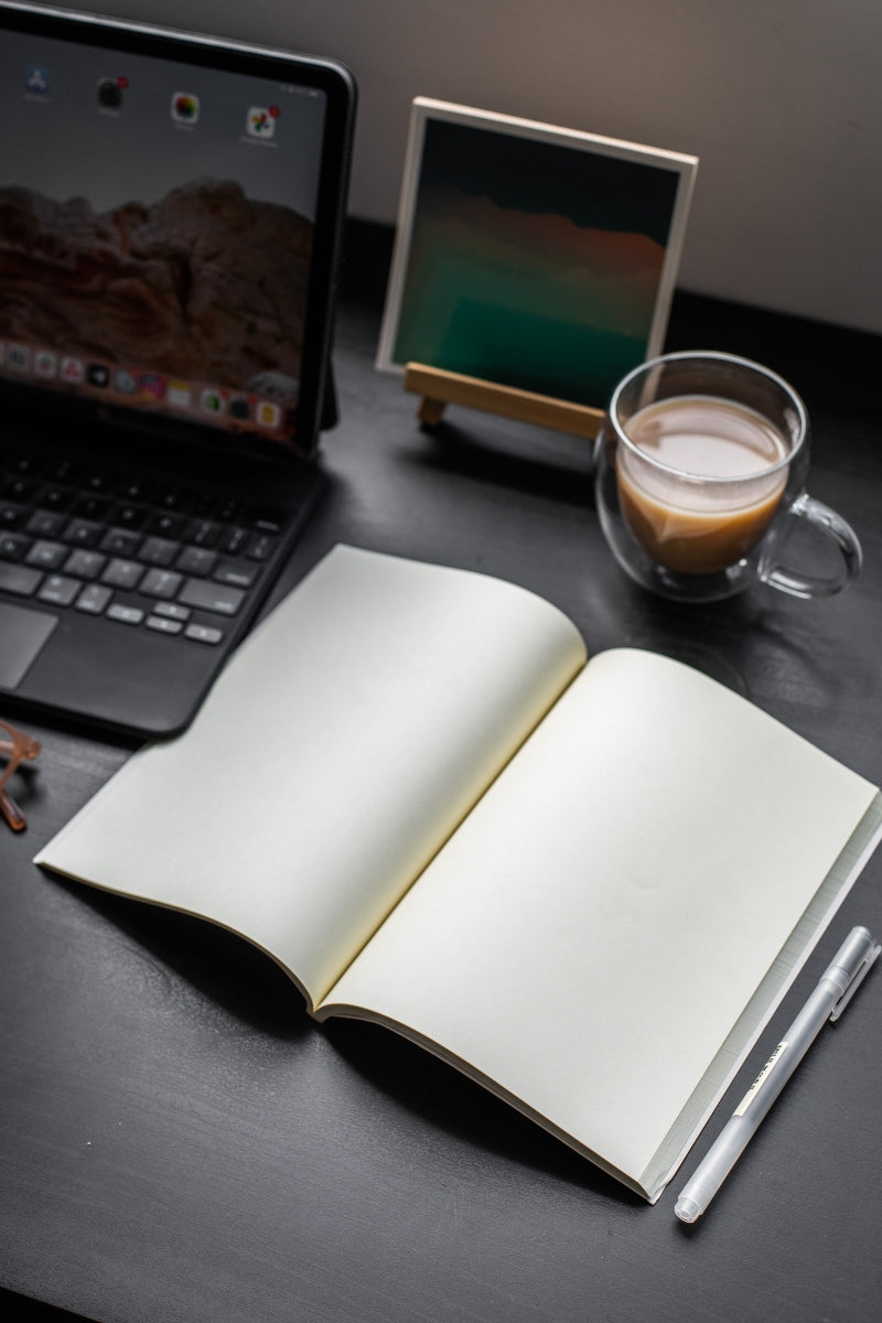 A blank, opened notebook laying on a black desk illuminated by soft sunlight and surrounded by a cup of coffee, a white pen, a iPad with a keyboard, and a picture frame.