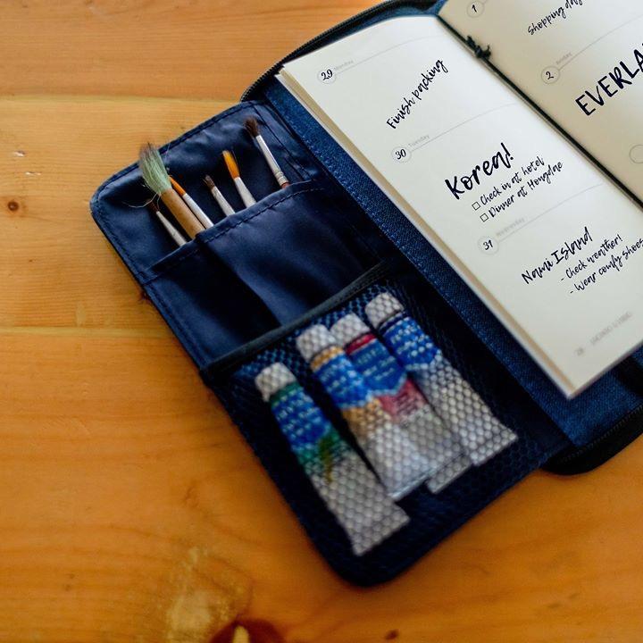A blue wallet that has in-built compartments that are being used to store paintbrushes and paint tubes.