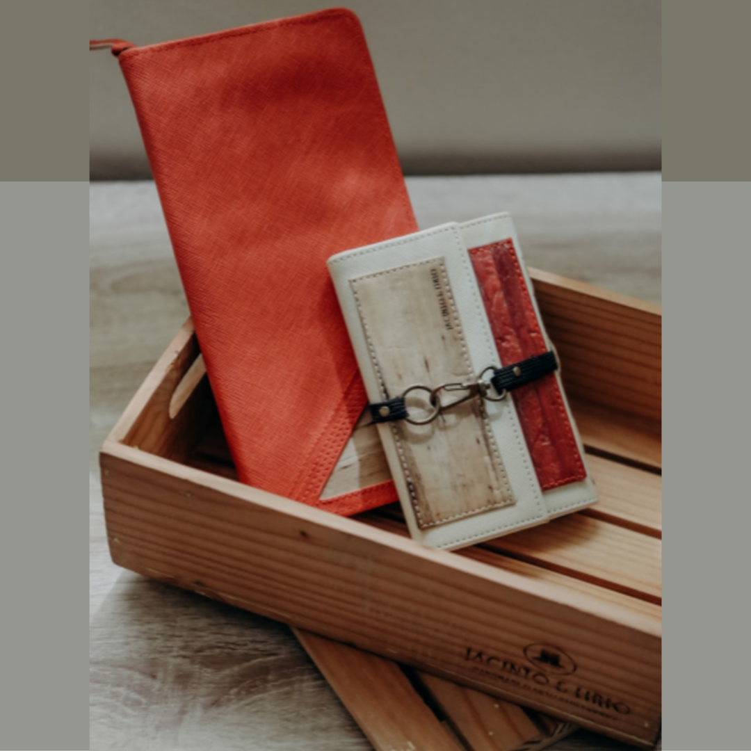 A souvenir gift set laying inside a small wooden crate. A rectangular orange vegan leather travel wallet and notebook and a white and red wallet with a metal hook closure.