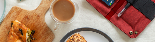 A cuttting board with a foccacia sandwich, a cup of coffee, and a red fabric tray with a pen.