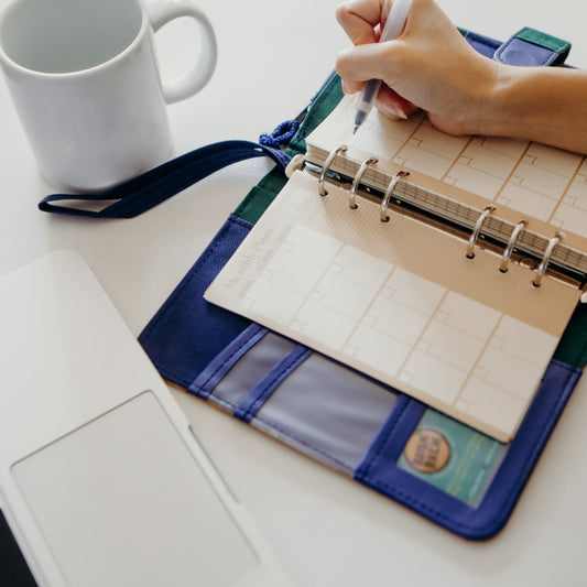 Person writing events on their planner, alongside a coffee mug and laptop.