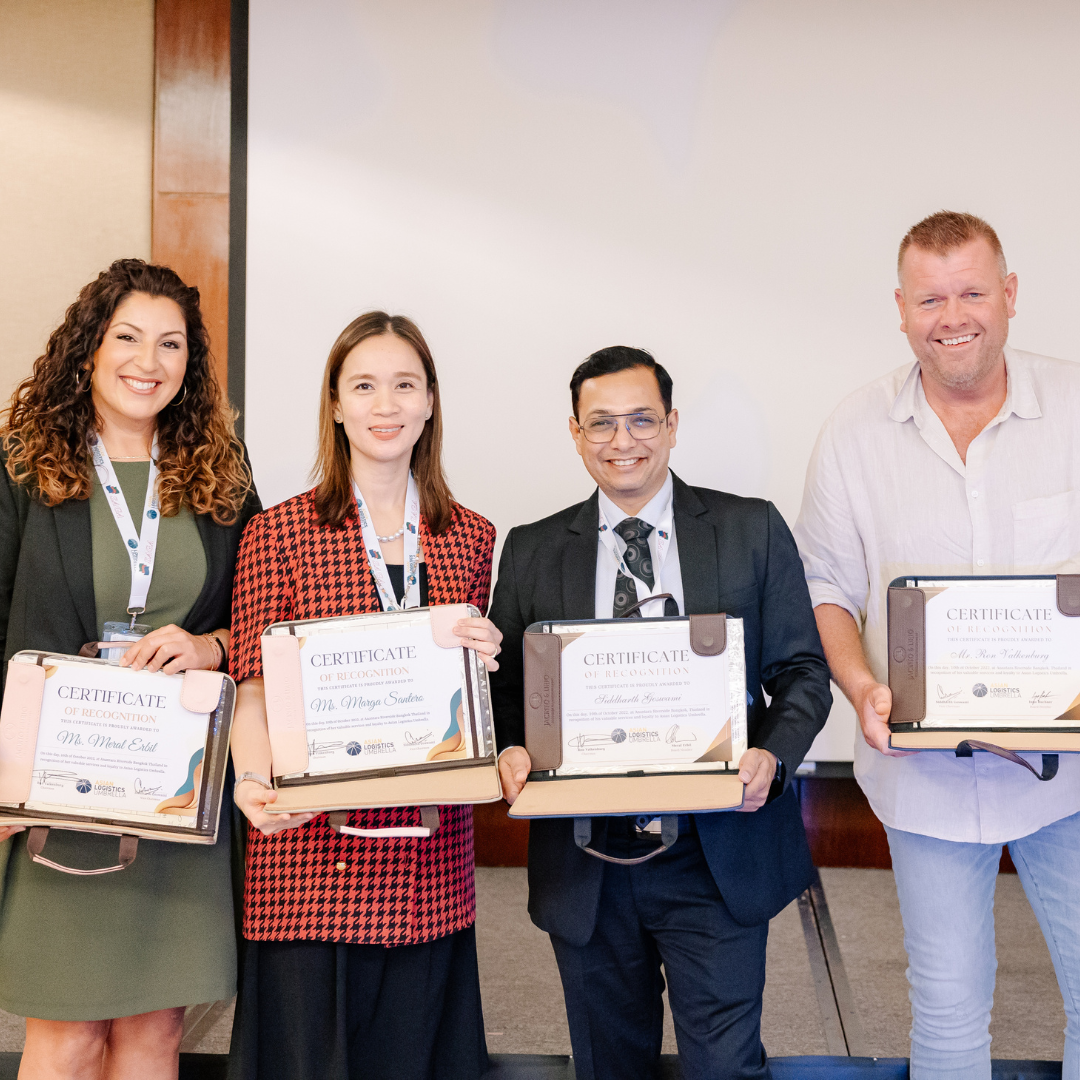 Four individuals standing side by side, each holding certificates of recognition in leather portfolio folders. The group includes two women and two men, all smiling, dressed in professional attire, with conference lanyards around their necks. The backdrop suggests a formal setting, likely an awards ceremony or business event.