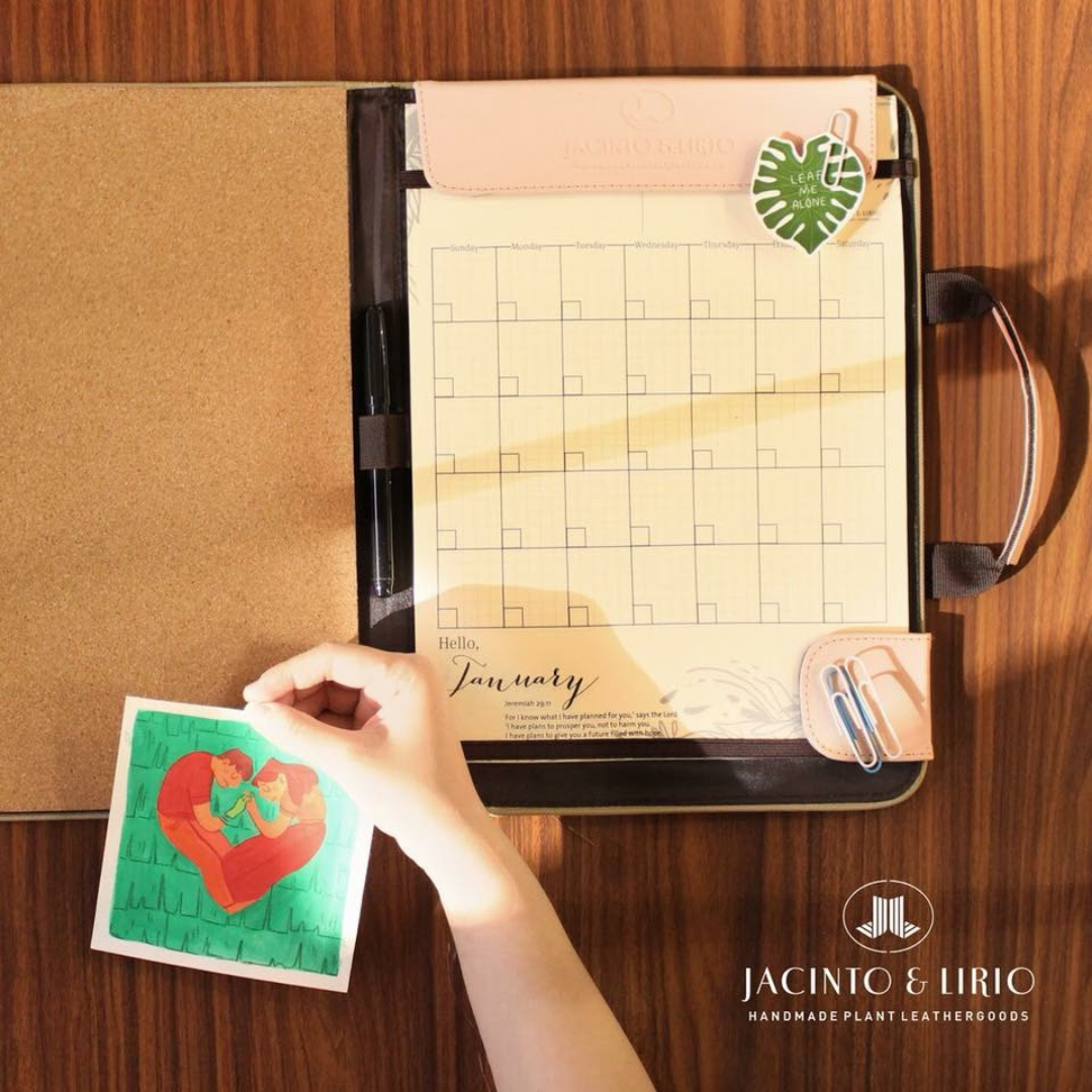 Open Jacinto & Lirio leather portfolio on a wooden desk, featuring a January calendar page and a corkboard on the left. A hand is holding a colorful illustration of two people embracing. The portfolio includes a leaf-shaped sticker, pens, and a sleek design, with the Jacinto & Lirio logo in the corner, promoting their handmade plant leather goods