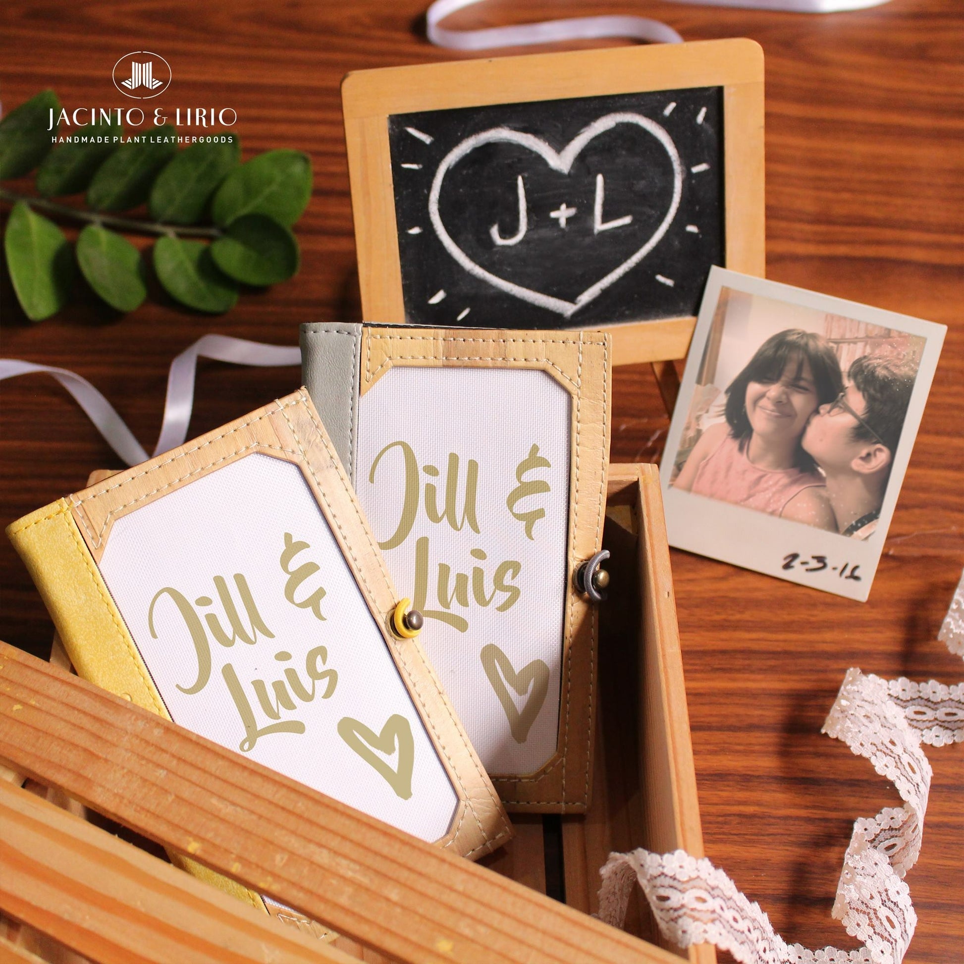 Two matching vegan leather journals in a wooden crate with the words "Jill & Luis" and a polaroid of a couple next to it