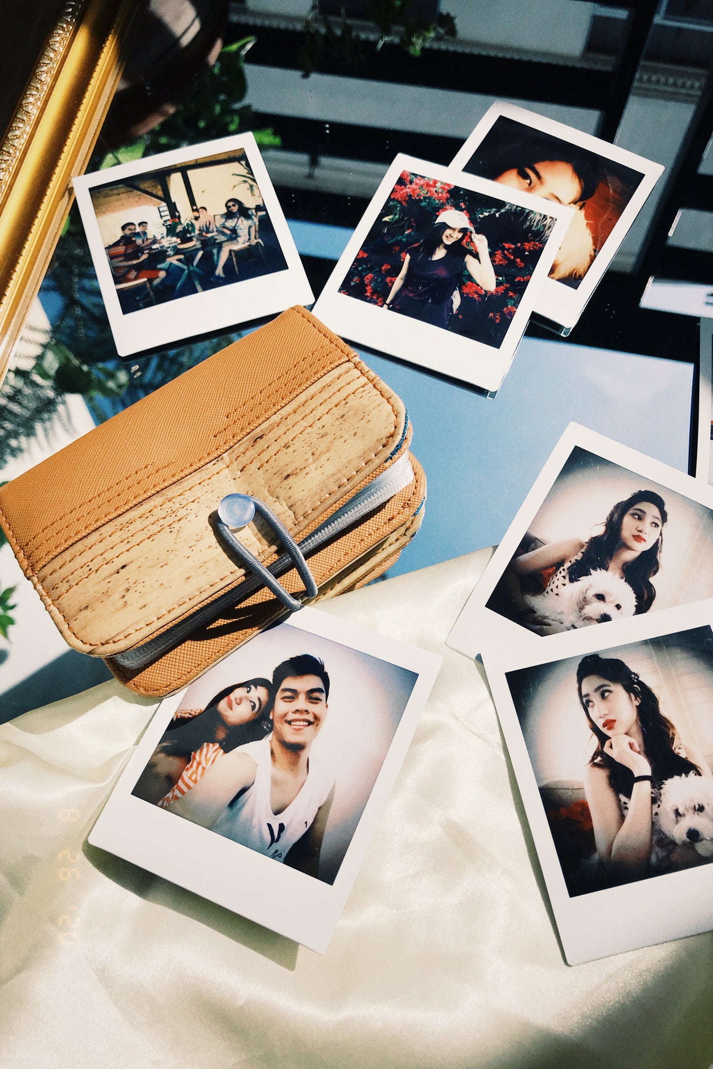 A light brown vegan leather wallet with a wooden-like exterior laying to the side of a mirror surface with polaroids of a couple scattered around it.