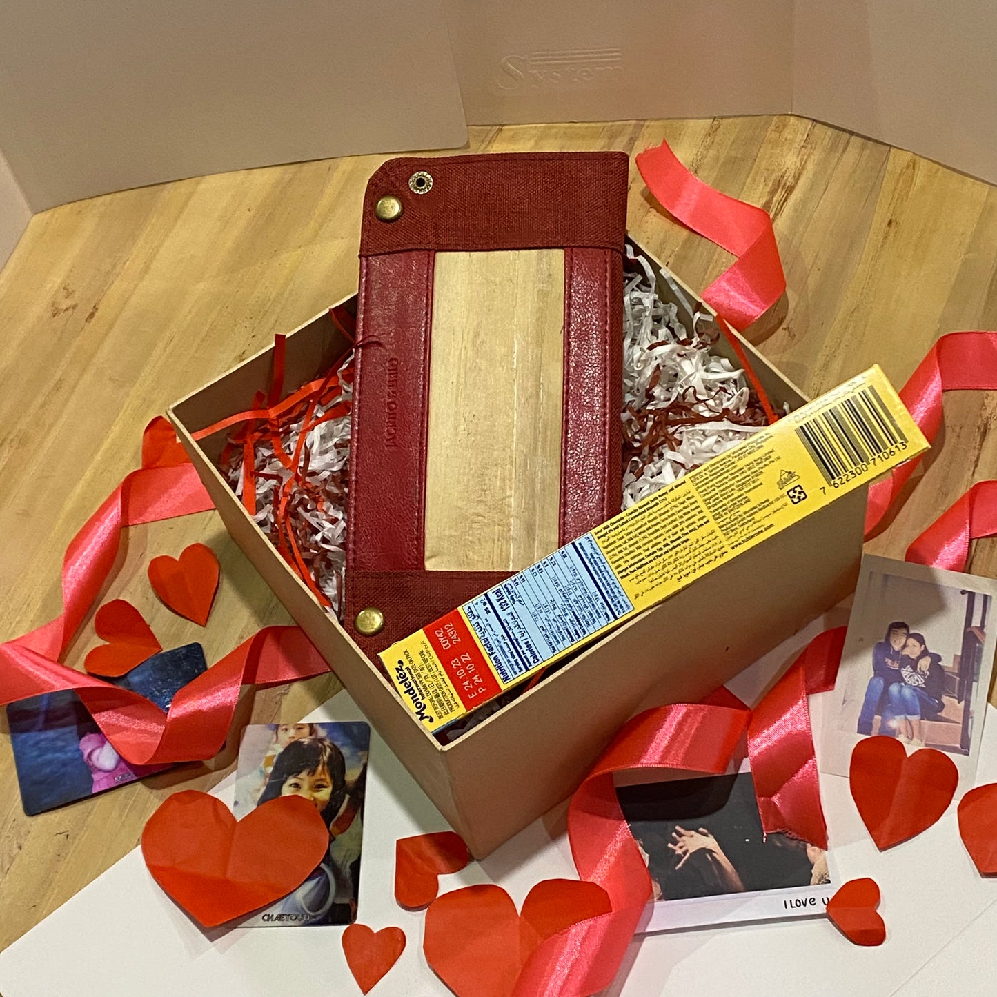 A valentines gift of a foldable vegan leather valet tray and a pack of Toblerone chocolate, laying in a cardboard box with red heart cutouts and twisted red ribbons strewn around it. 