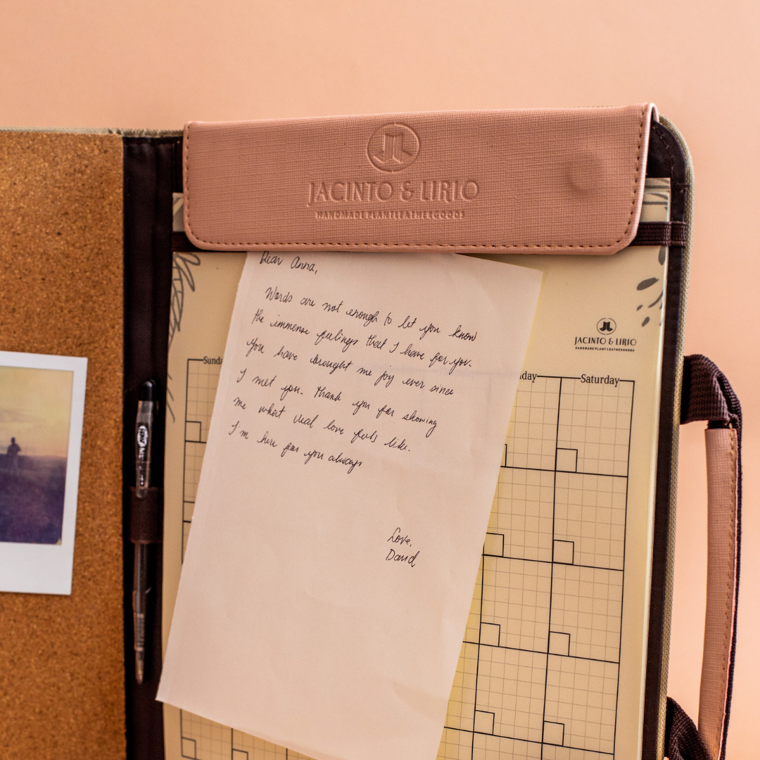 A Jacinto & Lirio leather organizer with a handwritten letter attached to the inside. The letter, addressed to 'Dear Doree,' expresses appreciation and gratitude. Behind the letter, a grid-style planner can be seen, along with a pen tucked into a holder. A small photo is pinned to the corkboard portion of the organizer. The scene suggests a personal, thoughtful message within a professional workspace setting.