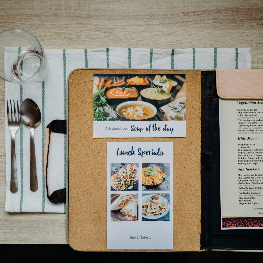 Restaurant table setting featuring a cork menu folder showcasing 'Soup of the Day' and 'Lunch Specials' promotions with vibrant food photos, including tacos, pasta, and soup. Next to the menu is a neatly arranged place setting with a fork, spoon, knife, napkin, and a glass, on a striped table runner. The menu highlights a 'Buy 1, Take 1' offer, adding an enticing dining option for guests.