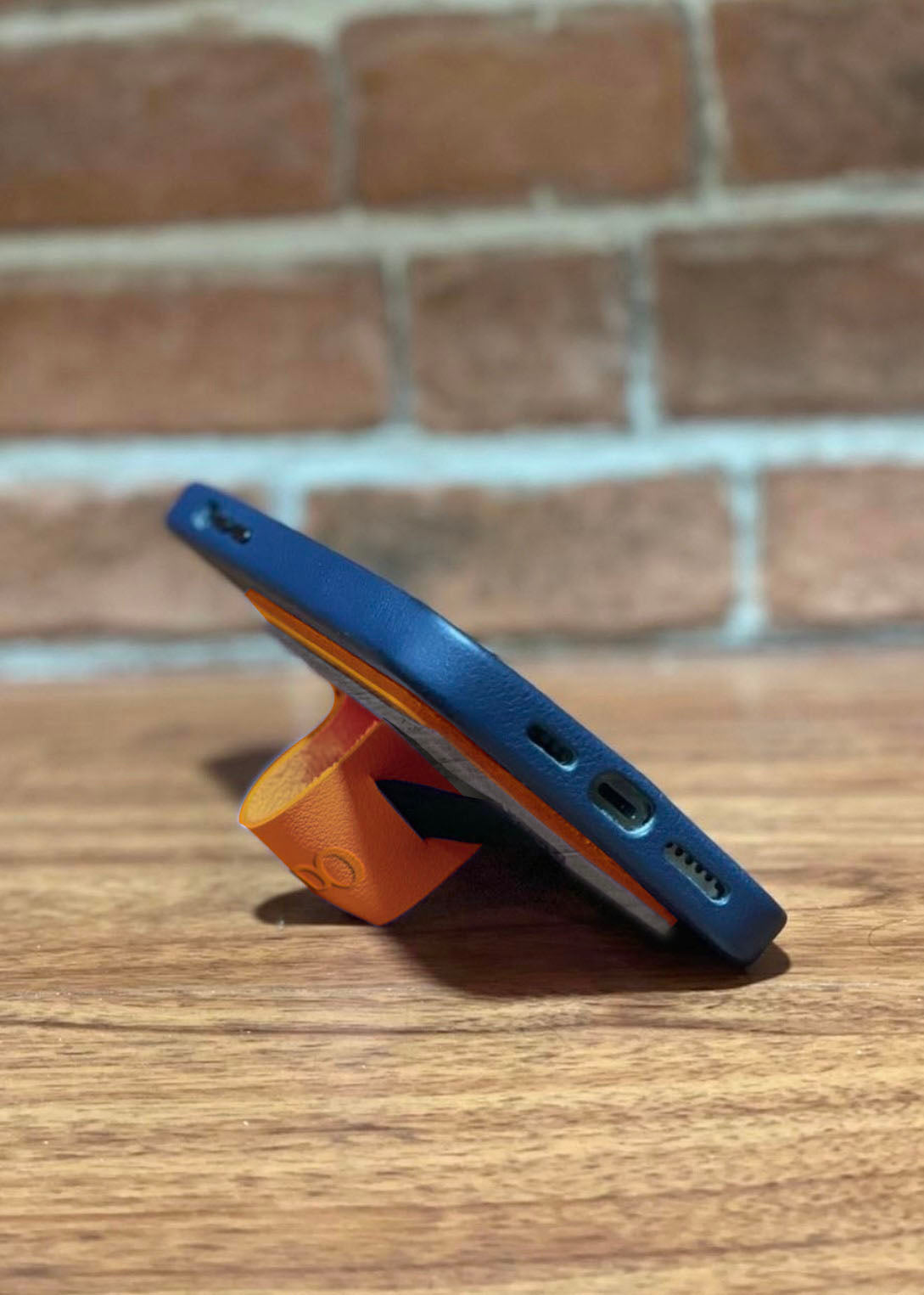 A side view of an orange vegan leather cellphone grip being used as a phone stand by folding the leather in on itself.