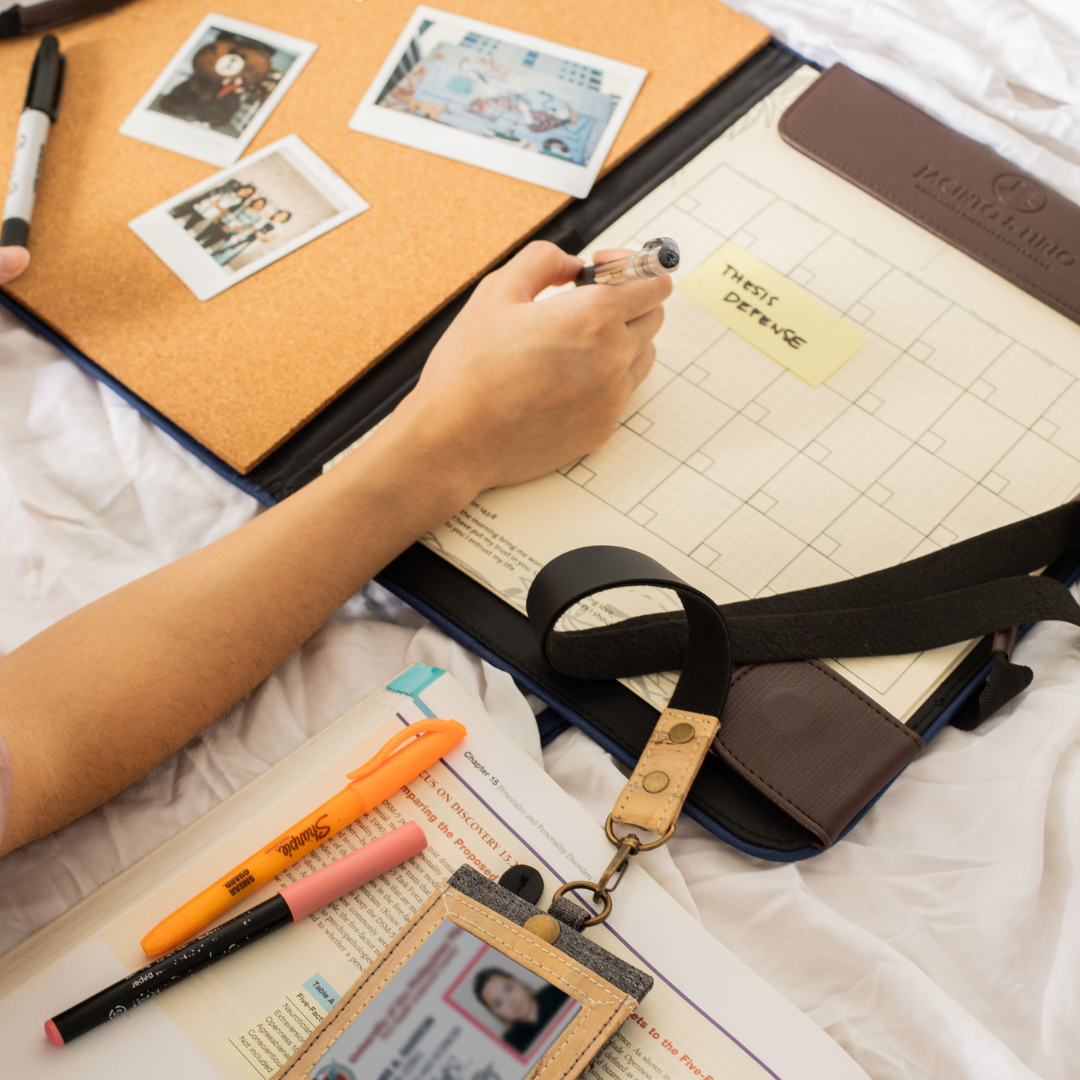 Open Jacinto & Lirio leather portfolio lying on a bed, with a hand holding a pen marking a planner with a sticky note that says 'Thesis Defense.' The portfolio’s corkboard side holds a few polaroid photos. Nearby are study materials, highlighters, and an ID badge, indicating a student preparing for an important academic event