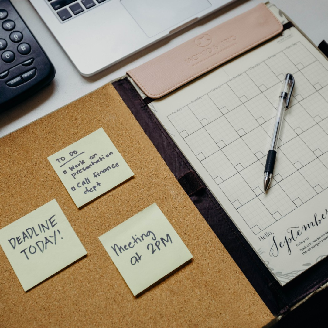 A workspace featuring a Jacinto & Lirio planner with a pen resting on a September calendar page. Beside it, a corkboard displays sticky notes with reminders, including 'Deadline Today!', 'Meeting at 2PM,' and a to-do list with tasks like 'Work on presentation' and 'Call finance dept.' A partial view of a laptop and office phone is visible, suggesting an organized, task-oriented office setting